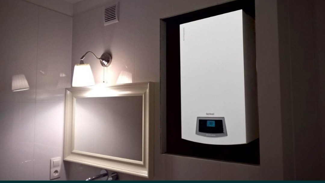 Nowoczesny, biały piec gazowy Termet zamontowany w niszy w łazience z białymi płytkami, obok lustra z oświetleniem.