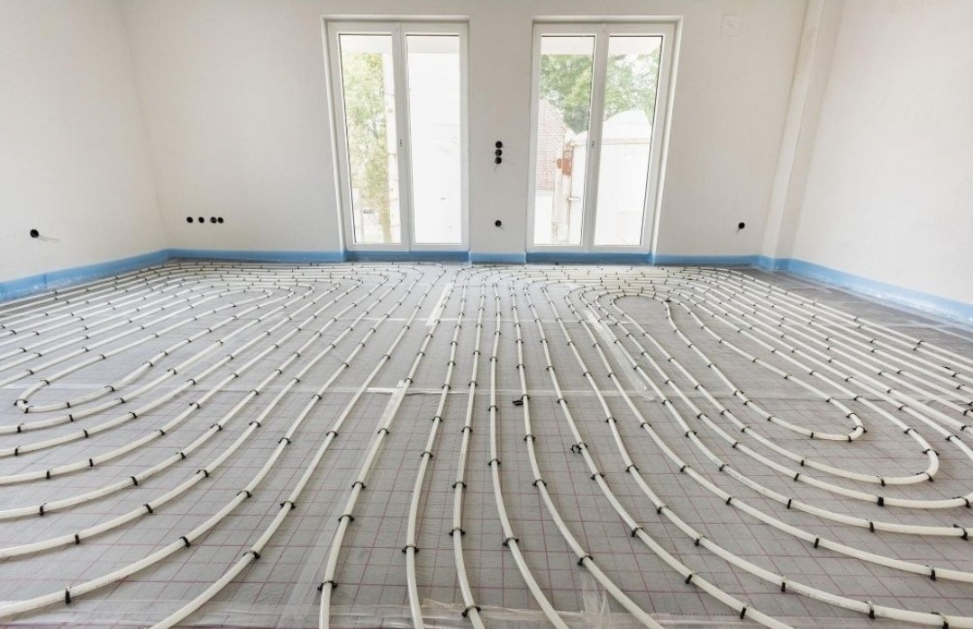 Białe rury ogrzewania podłogowego ułożone spiralnie na szarej macie izolacyjnej w jasnym pomieszczeniu z dwoma oknami i zabezpieczoną folią ścianą.