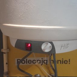 Paweł Bogusz - Bojler elektryczny marki Hit z widocznymi przyłączami, włącznikiem z czerwoną diodą i pokrętłem regulacji, zamontowany na żółtej ścianie z napisem 'Polecają mnie!'