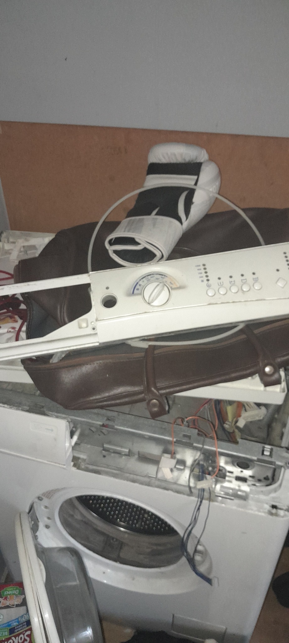 Rozłożona pralka automatyczna z widocznymi elementami wewnętrznymi, panelem sterowania i torbą skórzaną oraz rękawicą bokserską leżącymi na wierzchu.