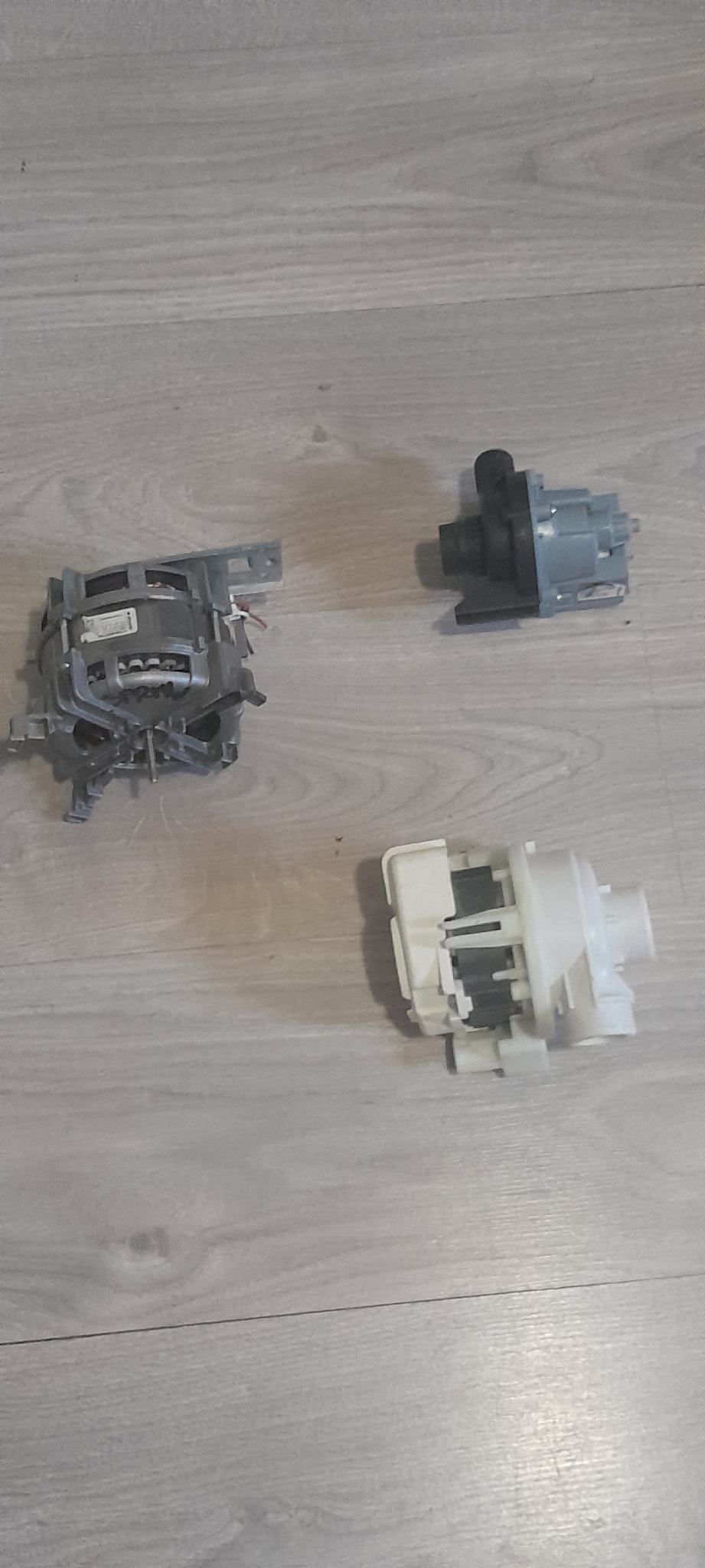 Silnik i dwie pompy do zmywarki lub pralki, leżące na panelach podłogowych o wzorze jasnego drewna.