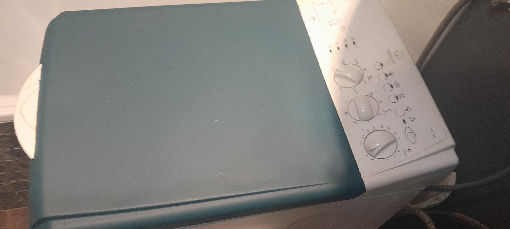 Pralka automatyczna Indesit WITL 105 z widocznym panelem sterowania z pokrętłami, sfotografowana z góry pod kątem, w tle fragment ściany i prawdopodobnie innej pralki.