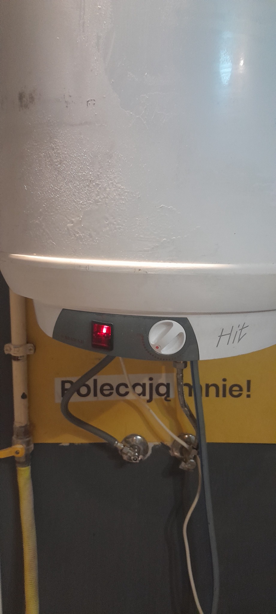 Bojler elektryczny marki Hit z widocznymi przyłączami, włącznikiem z czerwoną diodą i pokrętłem regulacji, zamontowany na żółtej ścianie z napisem 'Polecają mnie!'