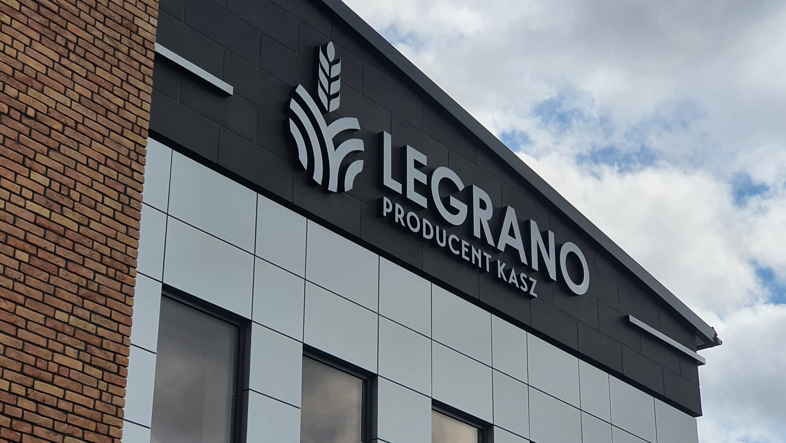 Elewacja budynku z logo firmy LEGRANO, producenta kasz, wykonanym w białych, trójwymiarowych literach na ciemnoszarym tle, z elementami graficznymi przedstawiającymi kłosy zboża. Widoczne fragmenty...