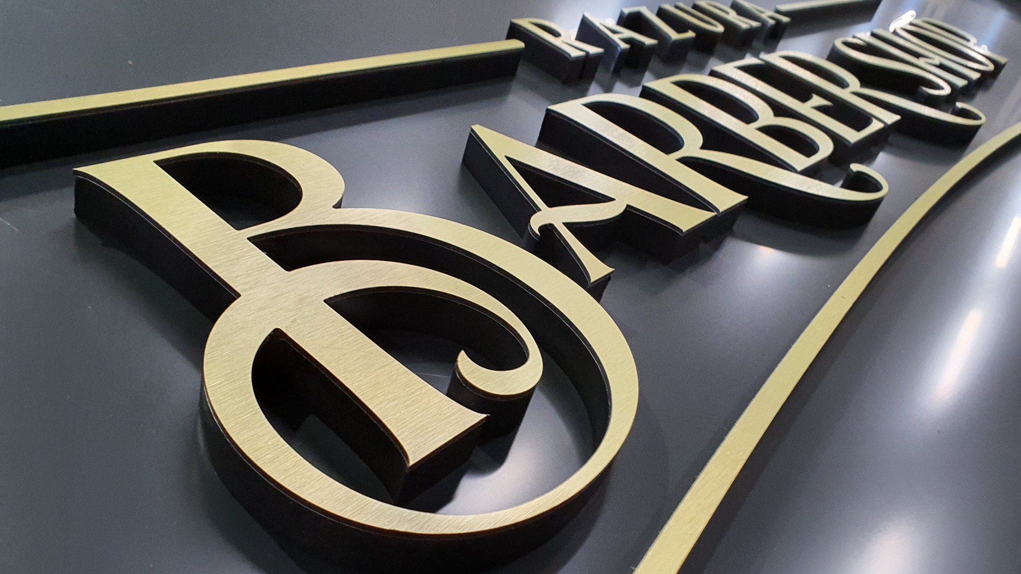 Elegancki szyld 3D z nazwą 'RAZURA BARBER SHOP' w odcieniach złota i czerni, wykonany z precyzją i dbałością o detale, prezentowany pod kątem.