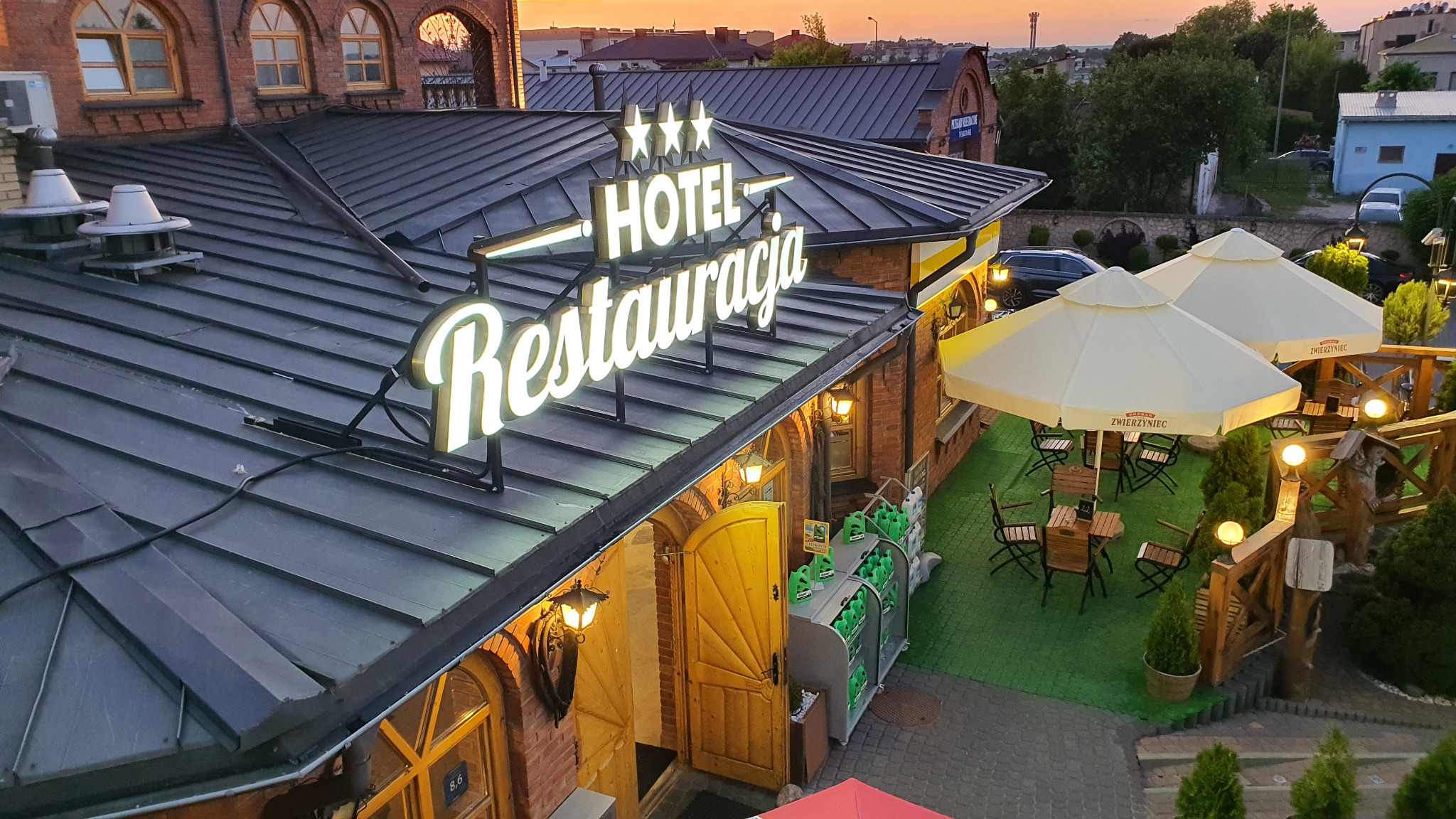 Widok z góry na oświetlony neonowy szyld 'Hotel Restauracja' z trzema gwiazdkami, umieszczony na dachu budynku z cegły, z widocznym ogródkiem restauracyjnym z parasolami i stolikami.