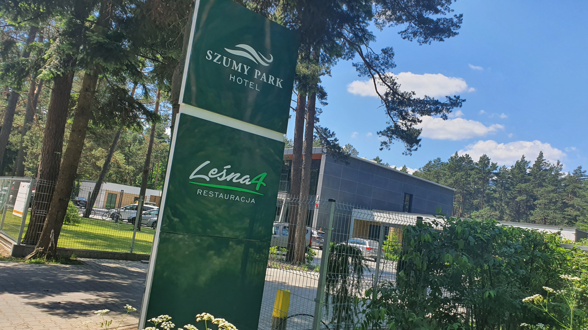 Zewnętrzny szyld reklamowy hotelu Szumy Park i restauracji Leśna 4, z widocznym budynkiem w tle, otoczony zielenią i drzewami.