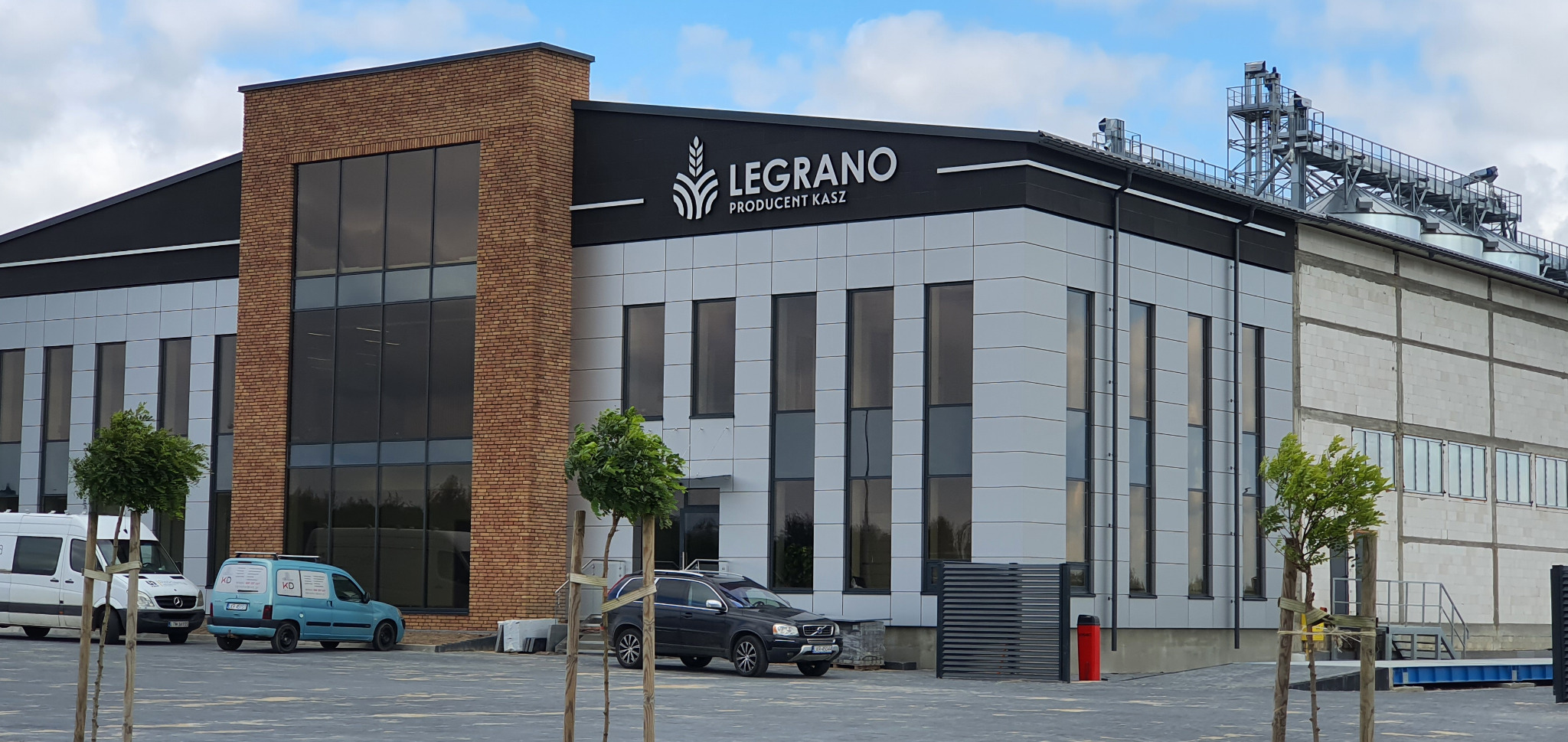 Nowoczesny budynek firmy Legrano, producenta kasz, z logo na elewacji, widoczne samochody dostawcze i osobowe na parkingu