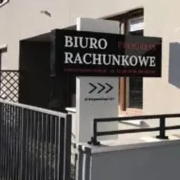 Czarny szyld biura rachunkowego z białymi napisami, umieszczony przed budynkiem z jasną elewacją. Pod szyldem strzałka wskazująca kierunek i adres.