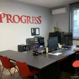 Nowoczesne biuro z dużym biurkiem, czerwonymi krzesłami i napisem 'PROGRESS' na ścianie, widok na okno z żaluzjami.