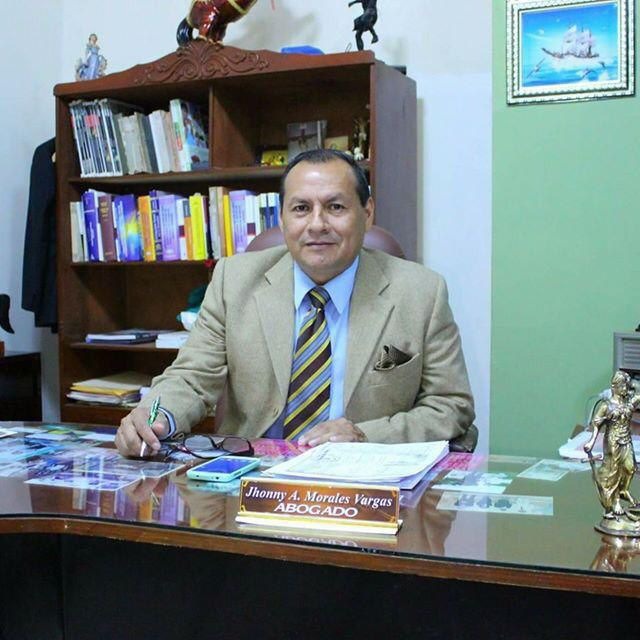Portret mężczyzny w beżowej marynarce i krawacie w paski, siedzącego za biurkiem z wizytówką 'Jhonny A. Morales Vargas ABOGADO', telefonem i okularami, w tle regał z książkami i obraz z żaglowcem.