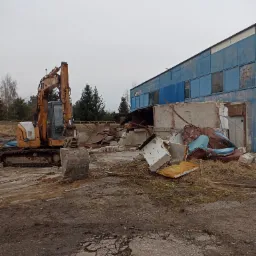 Pomarańczowa koparka gąsienicowa na placu budowy z gruzem i fragmentami zniszczonego budynku przemysłowego z niebieską elewacją w tle, widoczne uszkodzenia nawierzchni.