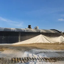 Budowa fundamentów: Widok na zaizolowany fundament zabezpieczony plandeką, z widocznymi materiałami budowlanymi na dachu budynku w tle i błękitnym niebem.