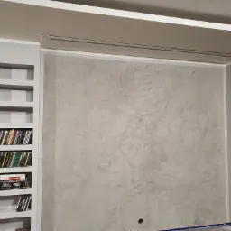 Ściana z szarą dekoracyjną fakturą tynku w ramie z półkami wnękowymi, wypełnionymi książkami i płytami CD.