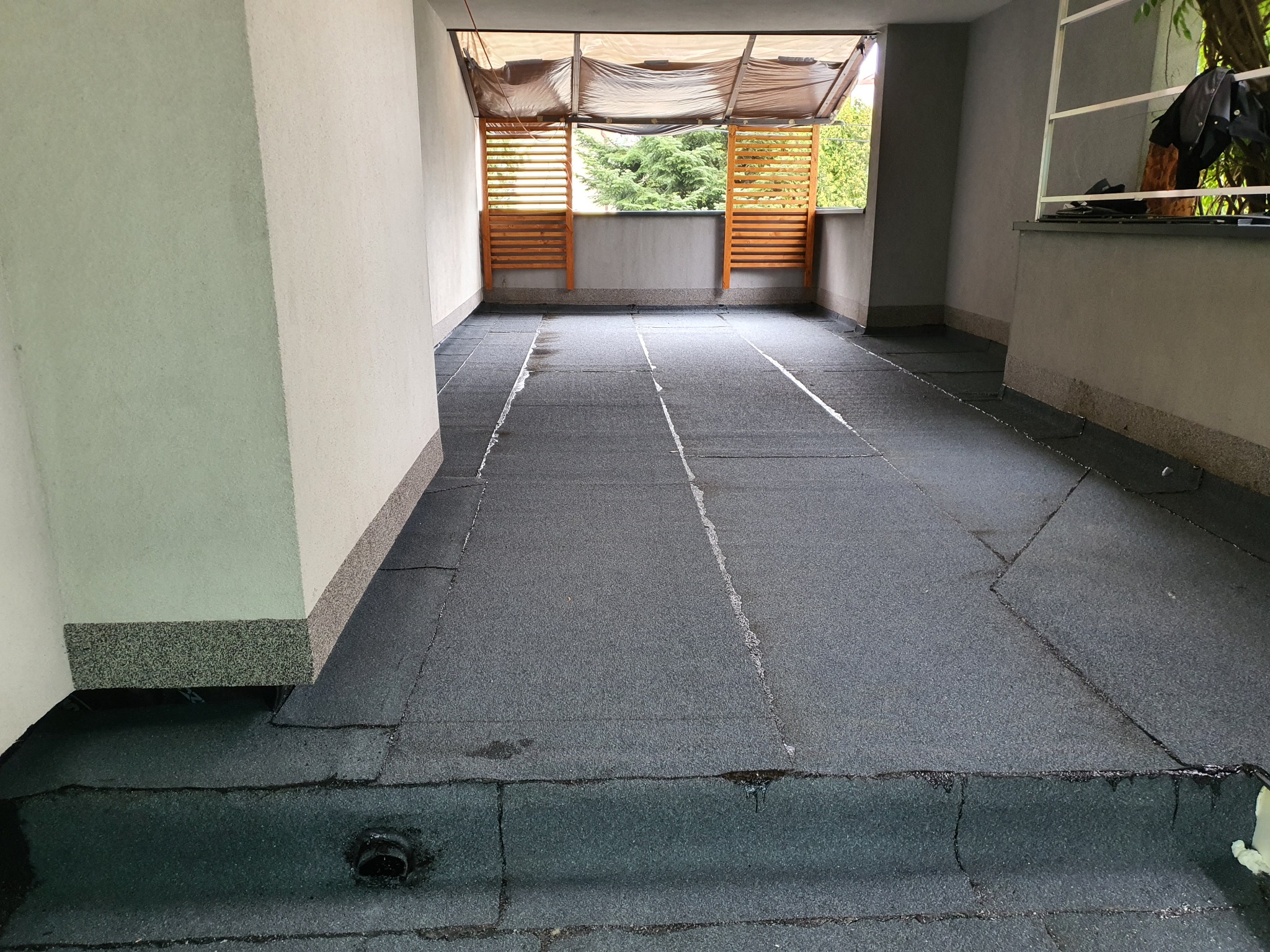 Balkon z nową, bitumiczną hydroizolacją, widoczne łączenia papy i odpływ. W tle drewniane panele balkonowe i zadaszenie z materiału.