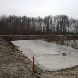 Świeżo wykopany staw z brudną wodą, widoczne ślady prac ziemnych, w tle rząd drzew bez liści i pochmurne niebo.