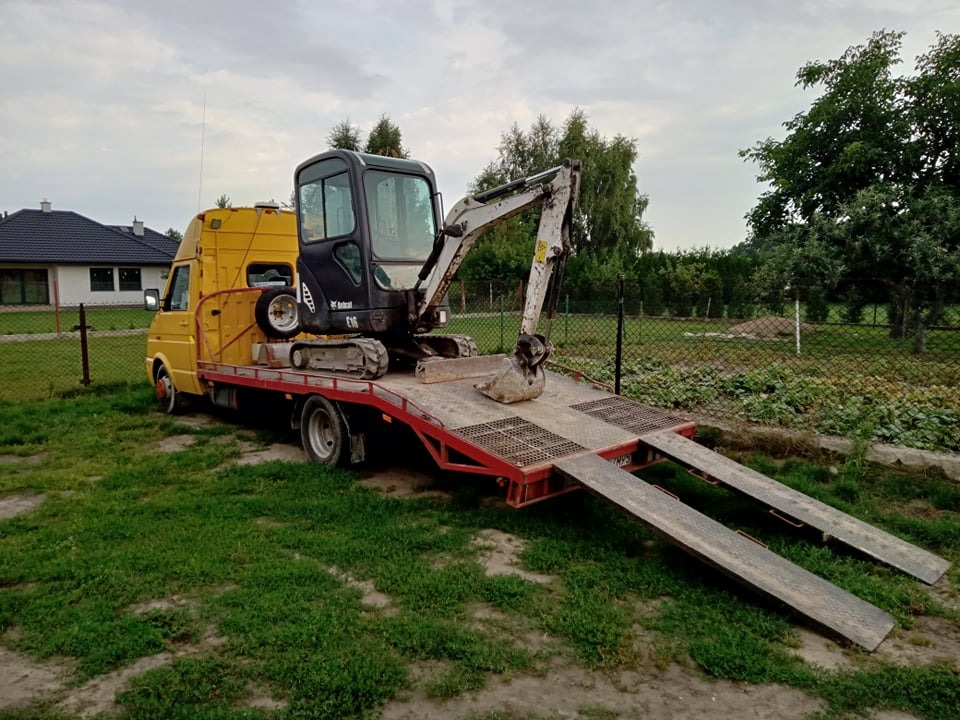 Żółty samochód dostawczy z platformą transportową przewożący małą koparkę Bobcat na tle zielonej trawy i ogrodzenia z siatki.