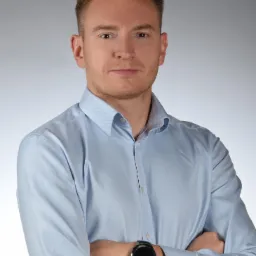 Portret młodego mężczyzny w błękitnej koszuli z zegarkiem na nadgarstku, skrzyżowane ręce, neutralne tło.