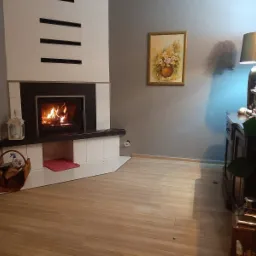 Nowoczesny kominek z białymi płytkami i czarnym obramowaniem, płomienie w palenisku, kosz z drewnem obok, drewniana podłoga, szare ściany z obrazem i komodą w tle.