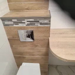 Wykończona toaleta z obudową stelaża wykonaną z płytek imitujących drewno, mozaiką i chromowanym przyciskiem spłukującym, widoczne białe płytki ścienne i marmurowe płytki podłogowe.