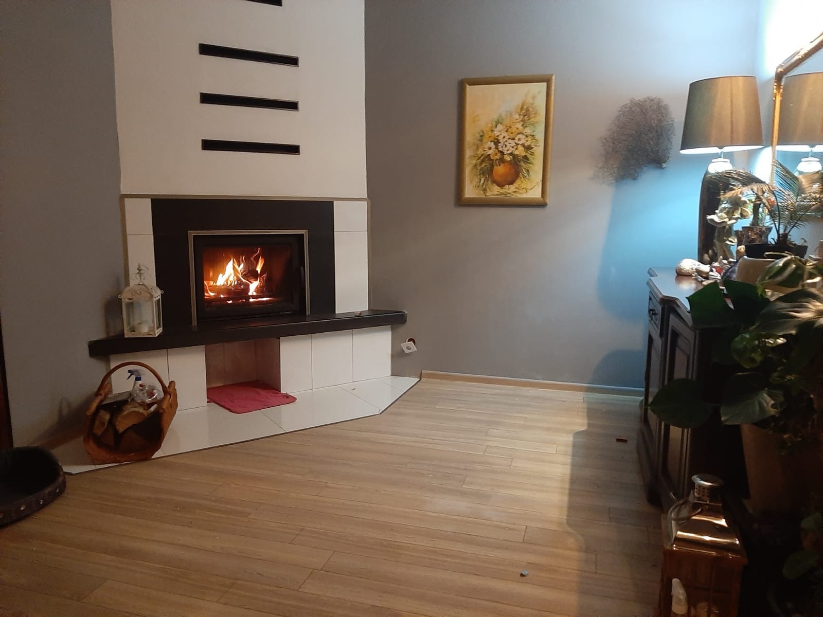 Nowoczesny kominek z białymi płytkami i czarnym obramowaniem, płomienie w palenisku, kosz z drewnem obok, drewniana podłoga, szare ściany z obrazem i komodą w tle.