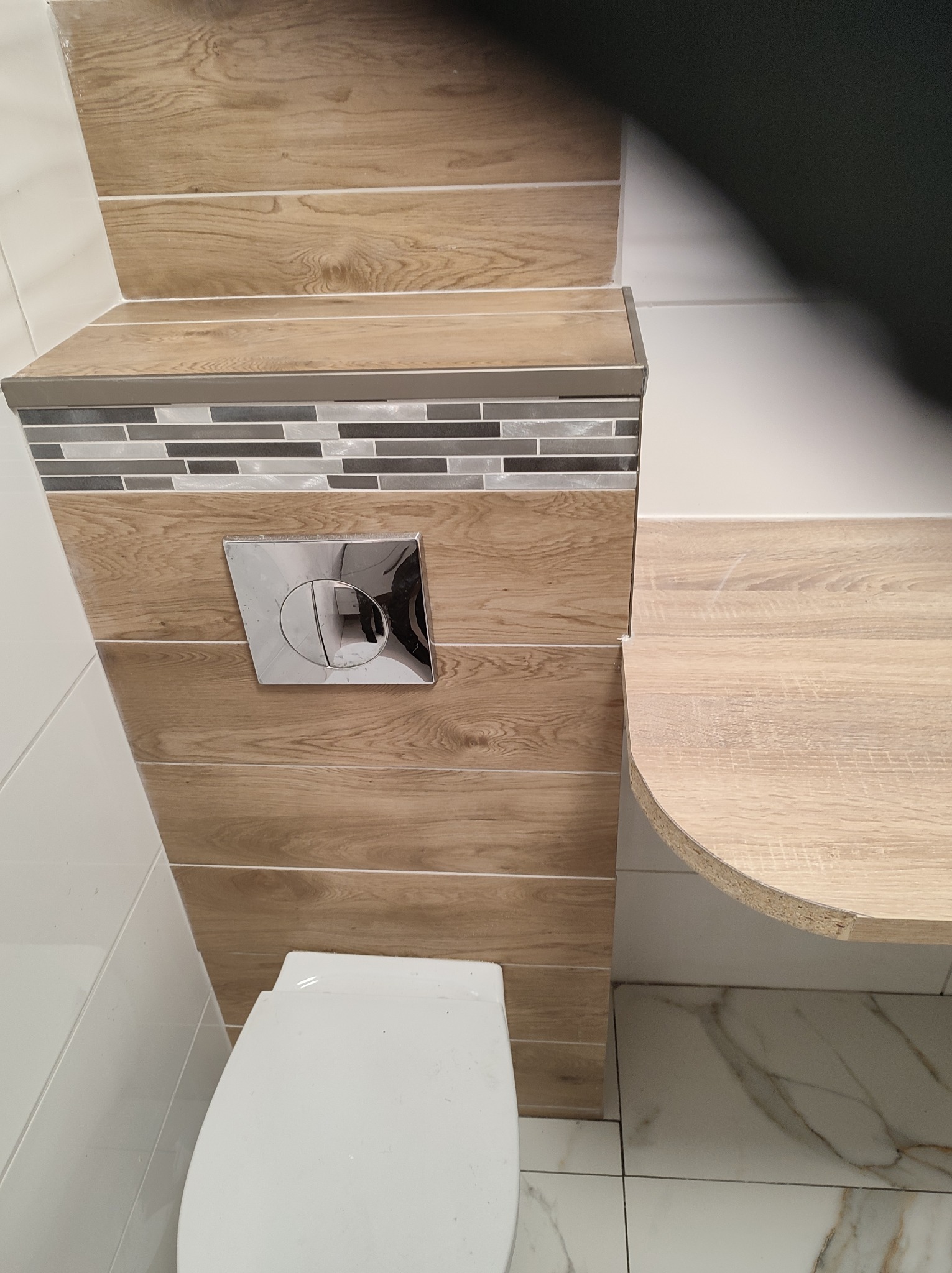 Wykończona toaleta z obudową stelaża wykonaną z płytek imitujących drewno, mozaiką i chromowanym przyciskiem spłukującym, widoczne białe płytki ścienne i marmurowe płytki podłogowe.