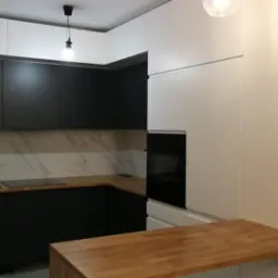 Nowoczesna kuchnia z czarnymi dolnymi szafkami i białymi górnymi, drewniany blat i marmurowy backsplash, oświetlenie punktowe.