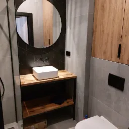 Nowoczesna łazienka z drewnianym blatem pod umywalką, czarną armaturą i geometrycznymi płytkami na ścianie, uzupełniona wiszącą szafką z drewnianymi frontami i podświetlanym lustrem.