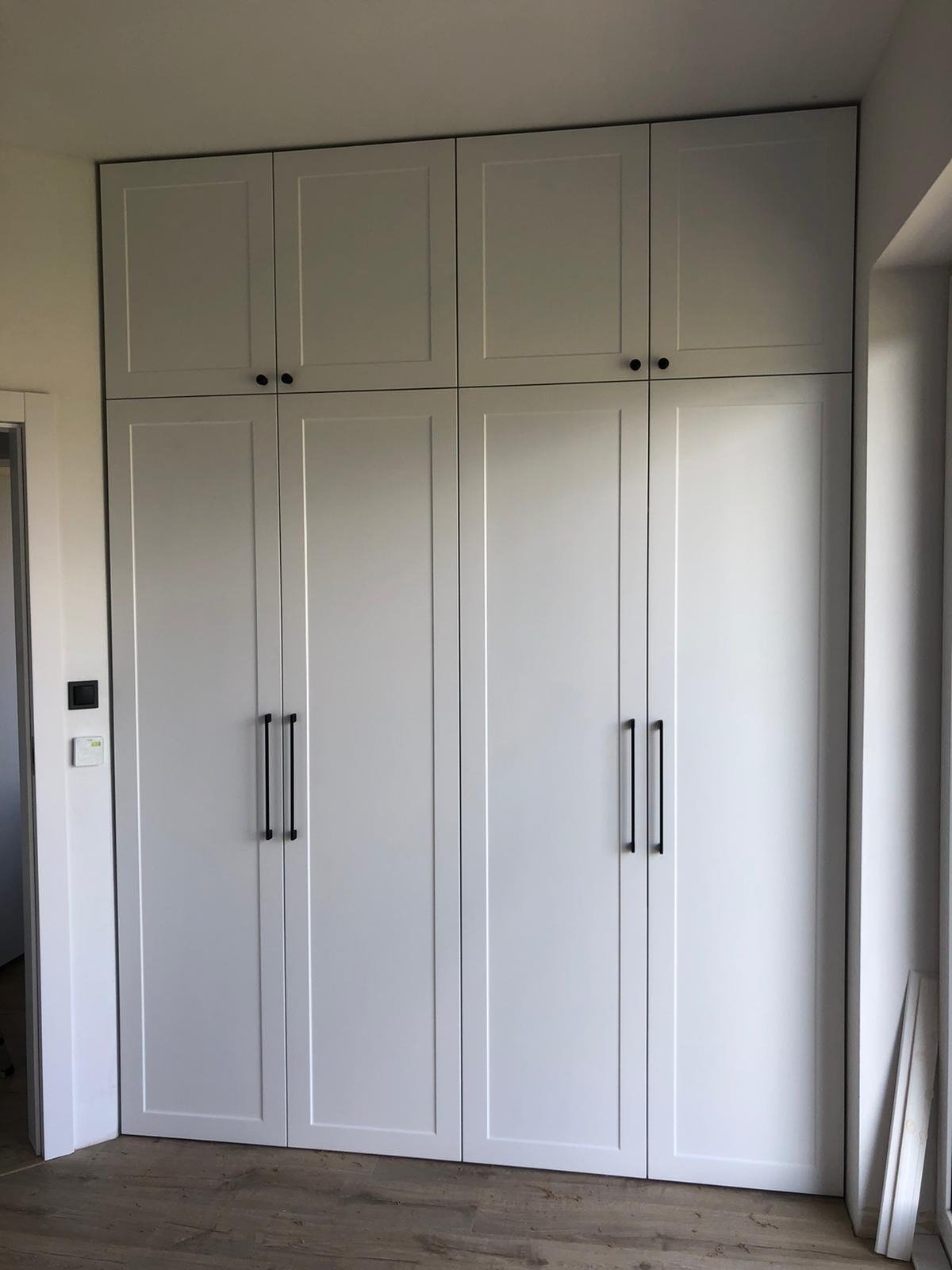 Biała szafa wnękowa z czarnymi, minimalistycznymi uchwytami, sięgająca sufitu, z dodatkowymi szafkami na górze, na podłodze panele imitujące drewno.