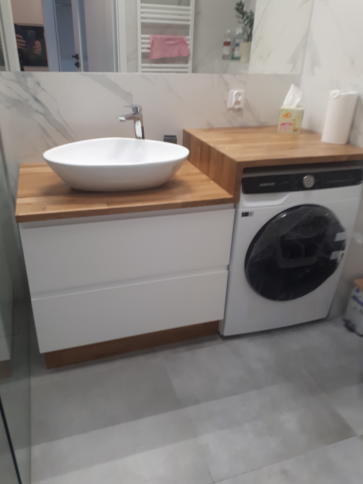 Nowoczesna łazienka z umywalką nablatową na szafce z białymi frontami i blatem z drewna, obok pralka Samsung z drewnianym blatem, białe płytki imitujące marmur na ścianie, grzejnik drabinkowy.