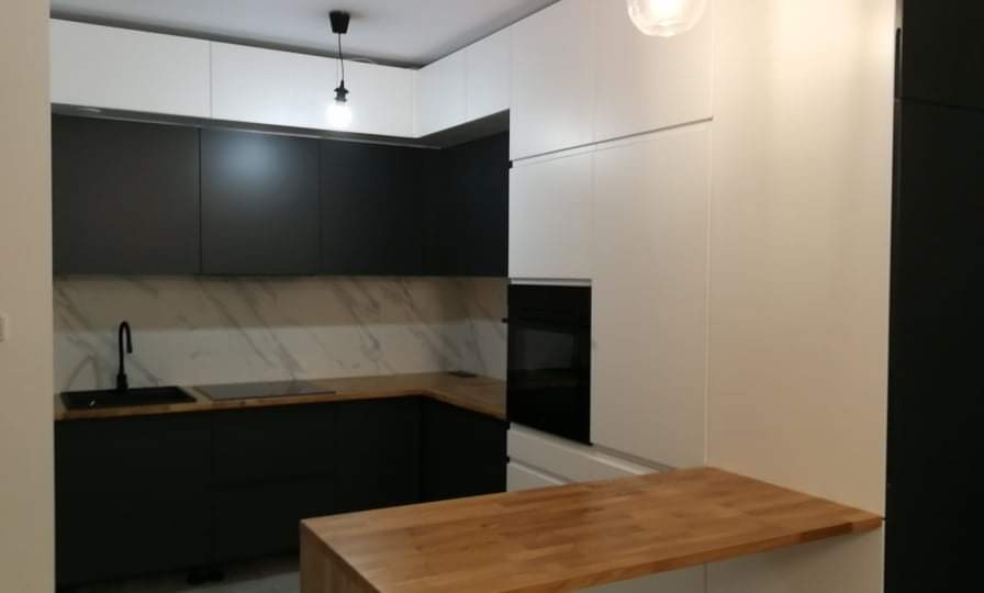 Nowoczesna kuchnia z czarnymi dolnymi szafkami i białymi górnymi, drewniany blat i marmurowy backsplash, oświetlenie punktowe.