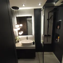 Nowoczesna łazienka w odcieniach szarości i czerni z podświetlanym lustrem, wiszącą umywalką, deszczownicą i dekoracyjnymi lampami wiszącymi.