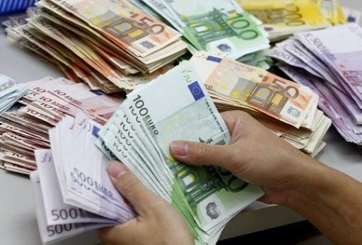 Dłonie liczą plik banknotów 100 euro na tle stosów banknotów o różnych nominałach, w tym 500, 150 i 100 euro.