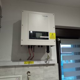 Lux Paweł Surowiak - Biały inwerter fotowoltaiczny marki SOFAR zamontowany na ścianie nad listwą z ostrzeżeniem o napięciu, widoczne przewody i roleta okienna w tle.