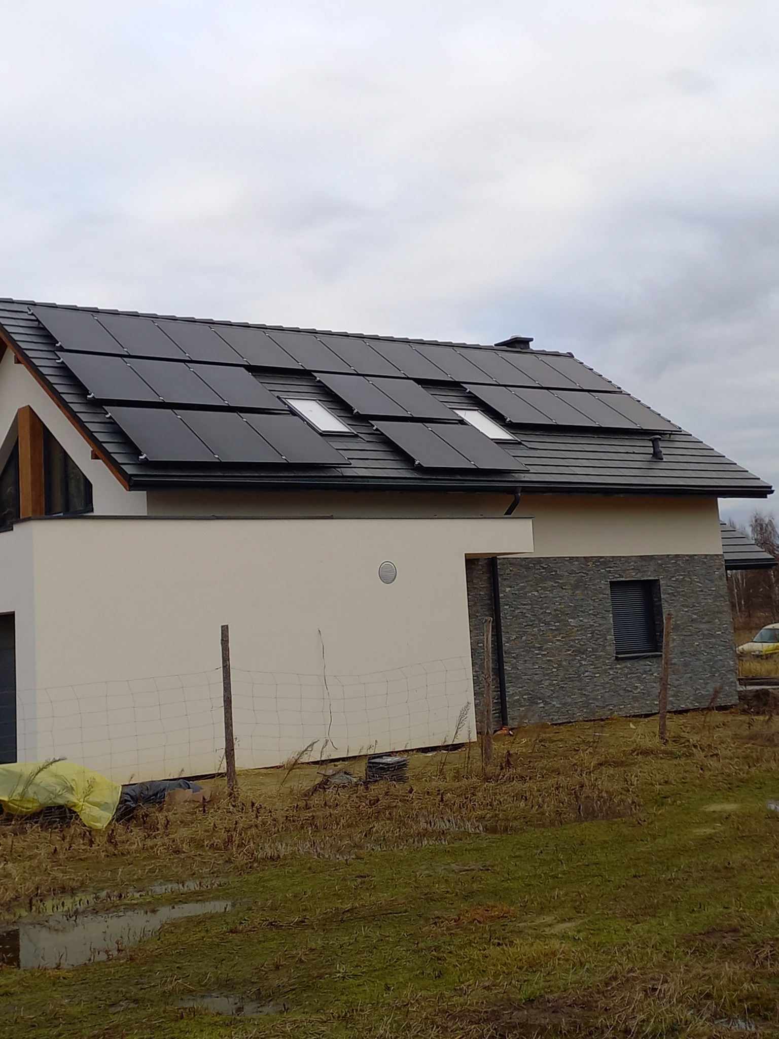 Nowoczesny dom jednorodzinny z panelami słonecznymi na dachu, widok z poziomu gruntu na podmokłą działkę, pochmurne niebo.