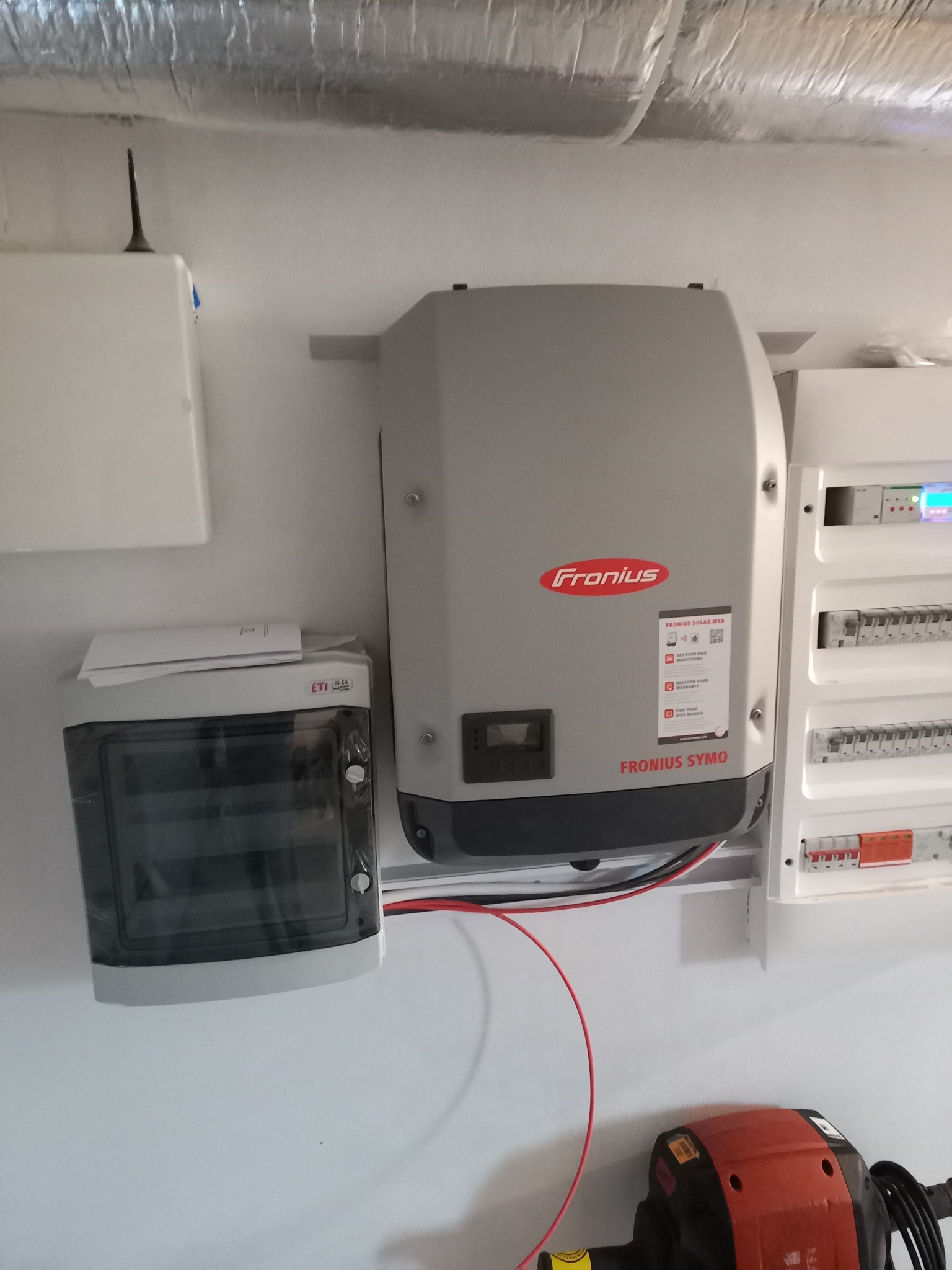 Szary inwerter Fronius Symo zamontowany na ścianie obok skrzynki elektrycznej z widocznymi przewodami i elementami instalacji fotowoltaicznej, z fragmentem pomarańczowego narzędzia na dole.