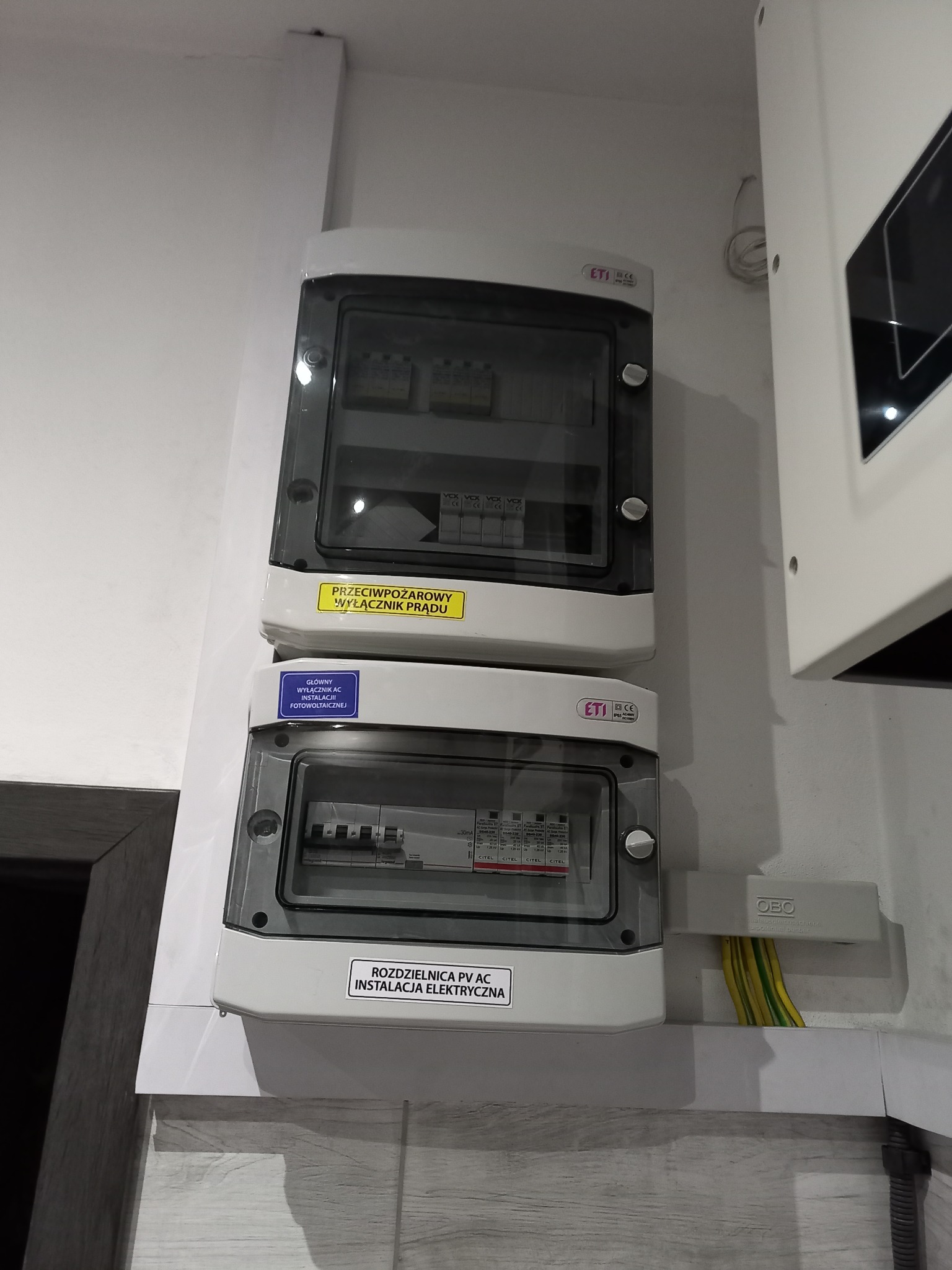 Dwa białe rozdzielnice elektryczne ETI z przezroczystymi drzwiczkami, górna z naklejką 'Przeciwpożarowy wyłącznik prądu', dolna z naklejką 'Rozdzielnica PV AC Instalacja elektryczna' i 'Główny...