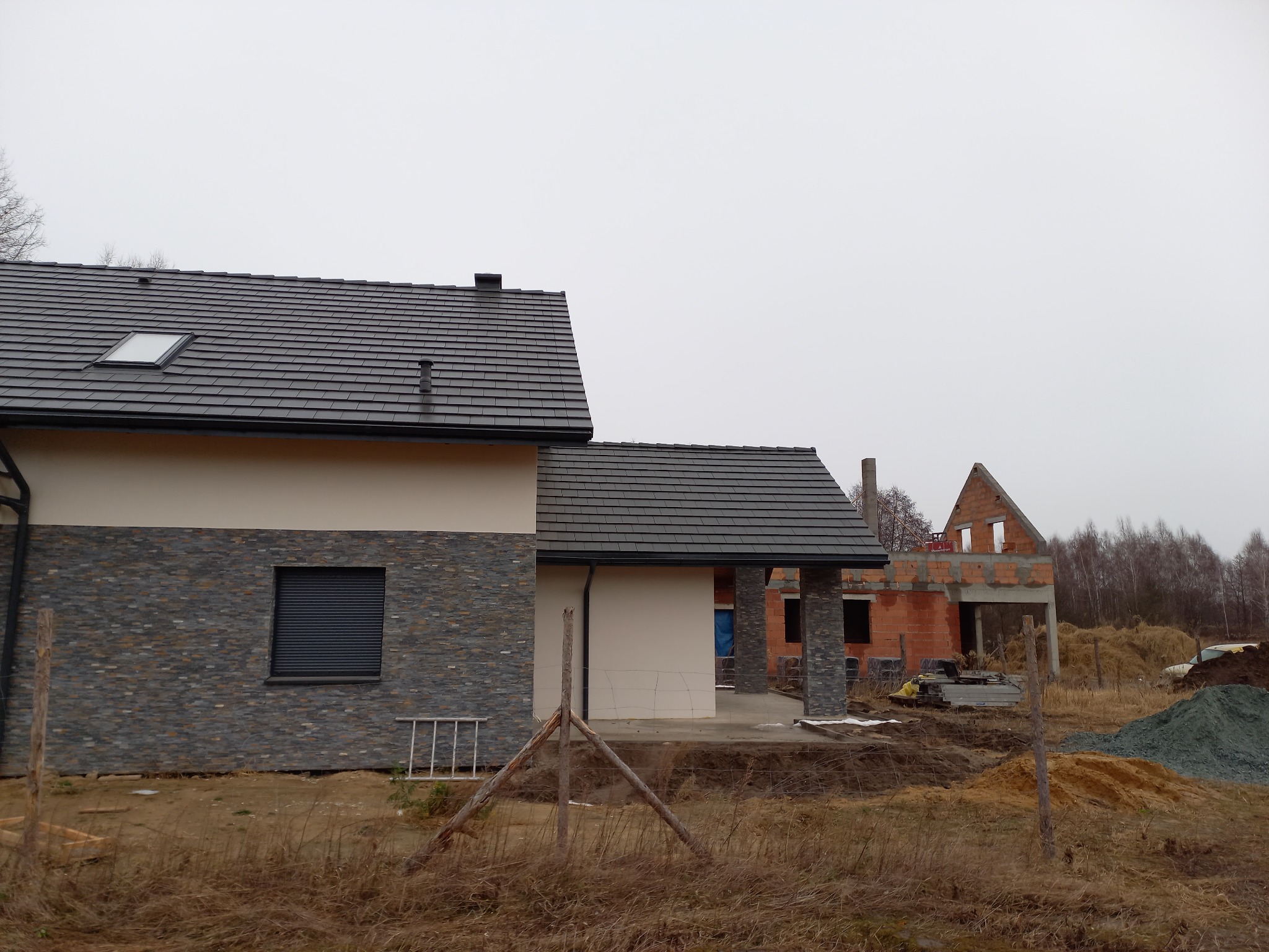 Dom jednorodzinny w trakcie budowy, z fragmentem elewacji wykończonej kamieniem dekoracyjnym, widoczny dach z oknem połaciowym i rusztowanie przy ścianie bocznej.