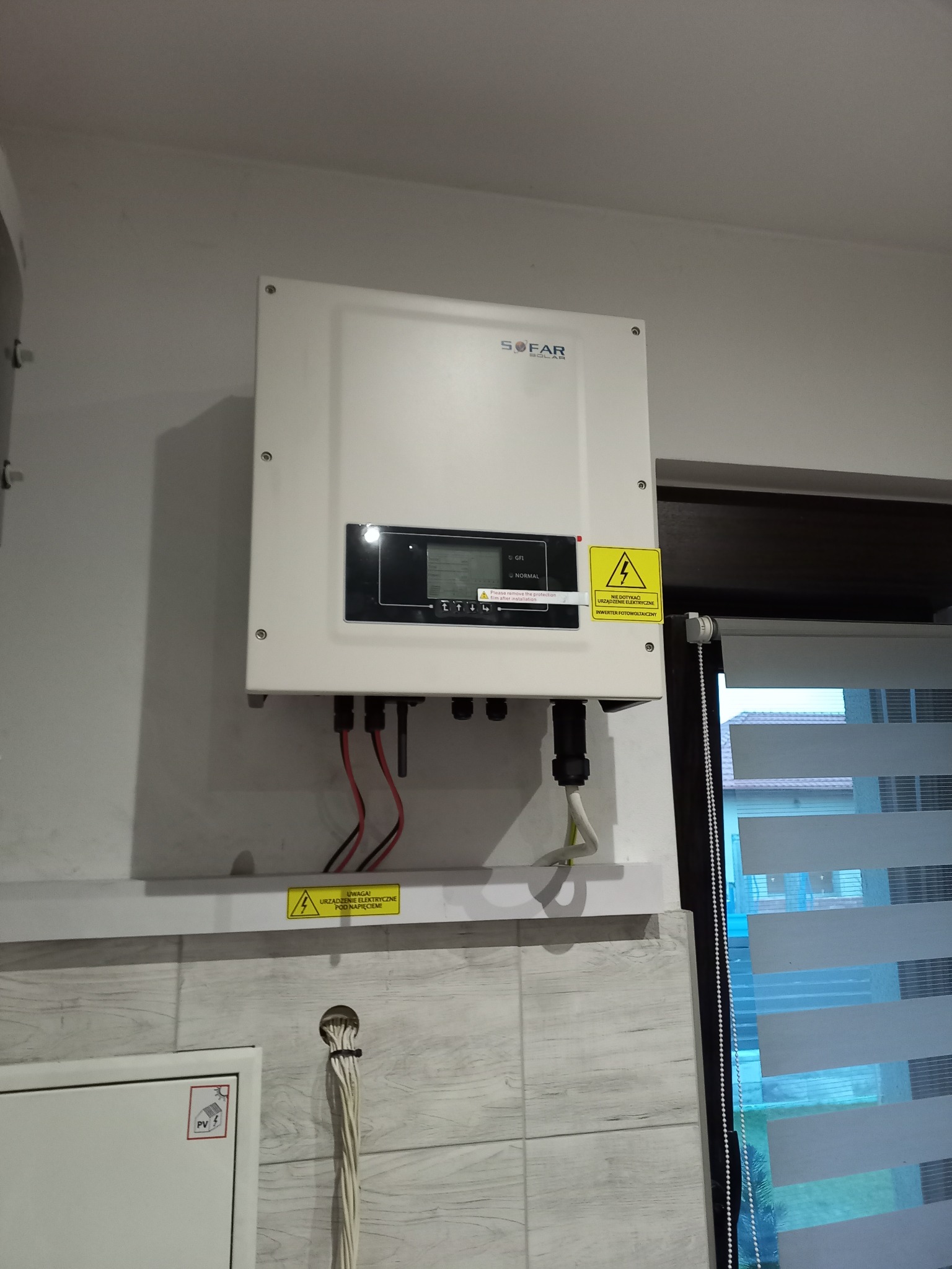 Biały inwerter fotowoltaiczny marki SOFAR zamontowany na ścianie nad listwą z ostrzeżeniem o napięciu, widoczne przewody i roleta okienna w tle.