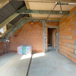 MAT-KAS Mateusz Kasperkiewicz - Surowy stan poddasza z instalacją elektryczną: przewody przymocowane do drewnianej konstrukcji dachu i ścian z cegły. Widoczne puszki instalacyjne i materiały budowlane.