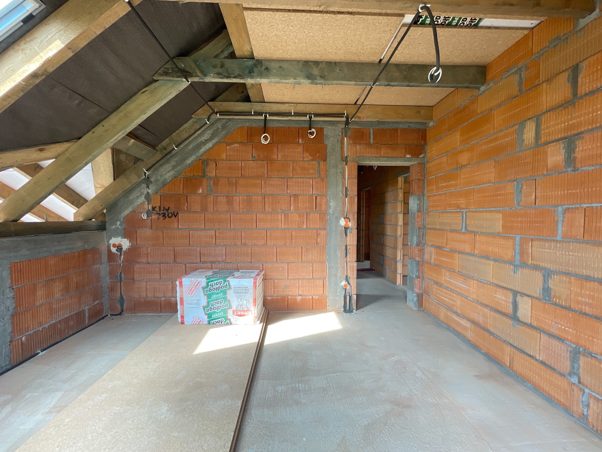 Surowy stan poddasza z instalacją elektryczną: przewody przymocowane do drewnianej konstrukcji dachu i ścian z cegły. Widoczne puszki instalacyjne i materiały budowlane.