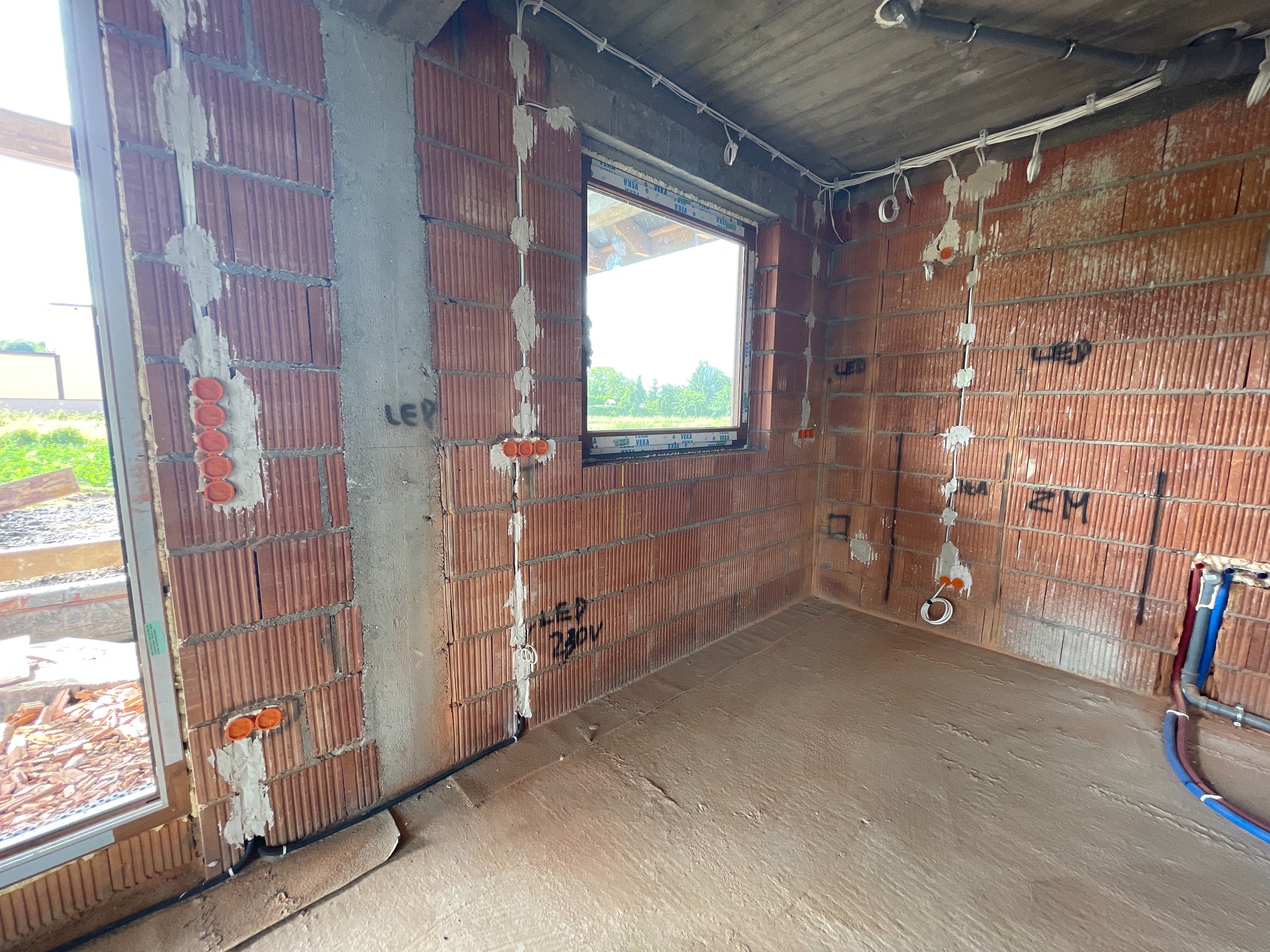 Surowy stan: instalacja elektryczna w nowym budynku z czerwonej cegły. Widoczne kable, puszki elektryczne i oznaczenia na ścianach. Podłoga wylewka.