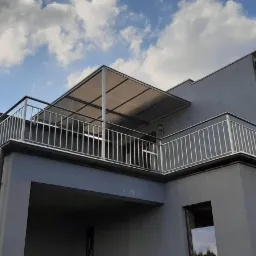Nowoczesny dom z szarą elewacją, balkon z metalową balustradą i zadaszeniem z poliwęglanu, widok z dołu na tle błękitnego nieba z chmurami.