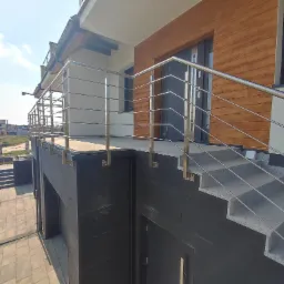 Zewnętrzne schody z szarego kamienia prowadzące do drzwi wejściowych, zabezpieczone minimalistyczną, metalową barierką z poziomych prętów, na tle budynku z drewnianą elewacją i widokiem na plac...