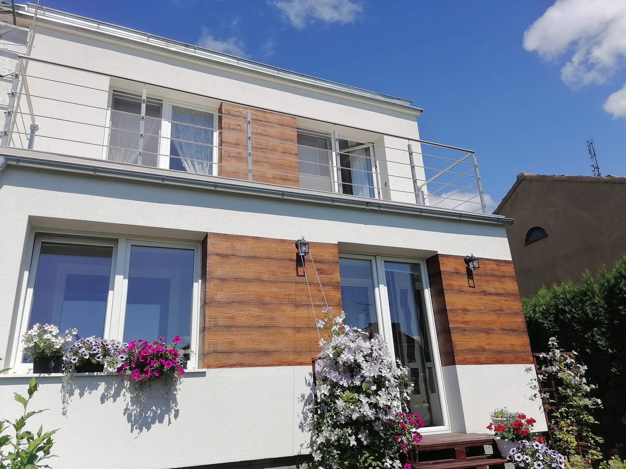 Nowoczesny dom z białymi ścianami, drewnianymi panelami, balkonem z metalową balustradą i kwitnącymi kwiatami w donicach na parapecie i przy wejściu.