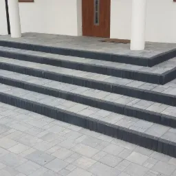 Schody wejściowe do domu wyłożone szarą kostką brukową z ciemnoszarym obrzeżem, widok z dołu, trawnik w tle.