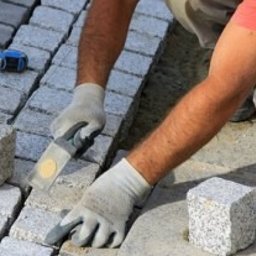 Bruk Bydgoszcz Usługi Brukarskie - Pracownik w rękawicach roboczych układa kostkę granitową, wyrównując ją gumowym młotkiem. Widoczny fragment chodnika z szarej kostki oraz narzędzia brukarskie.