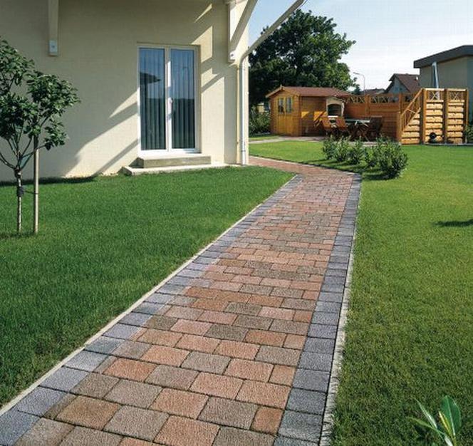 Kręta ścieżka z kostki brukowej w odcieniach brązu i szarości prowadząca przez zielony trawnik, obok domu z białymi drzwiami balkonowymi i drewnianej altany w tle.