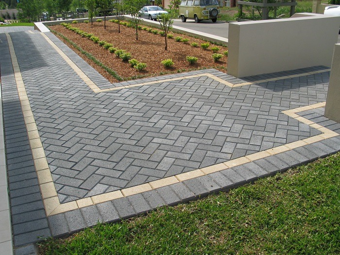 Szara nawierzchnia ułożona w jodełkę z obramowaniem z jasnych kostek, widok na podjazd i rabatę z młodymi drzewkami.