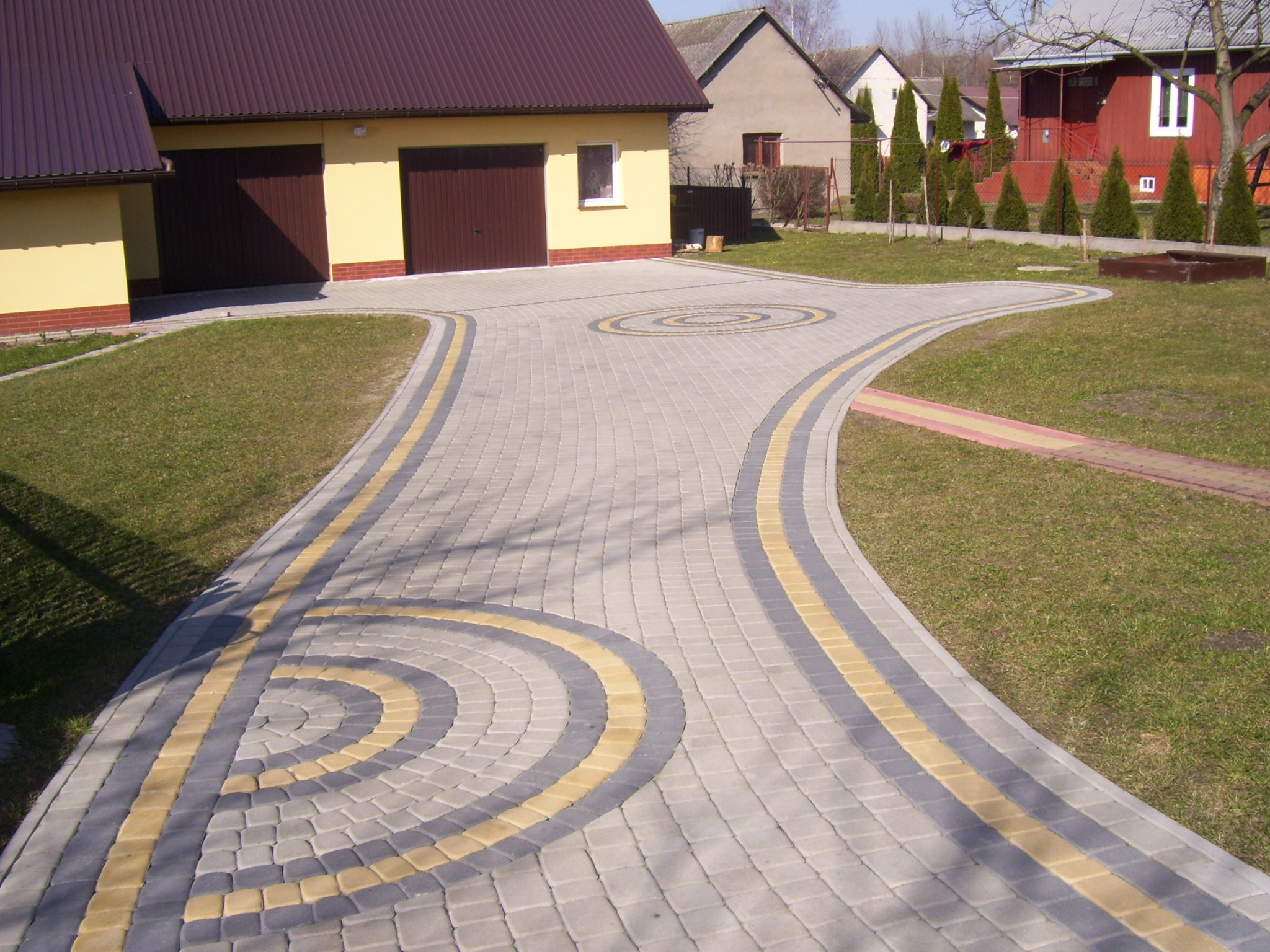 Szeroki, rozwidlający się chodnik z szarej kostki z żółtymi akcentami, prowadzący do garażu i domu, otoczony trawnikiem.